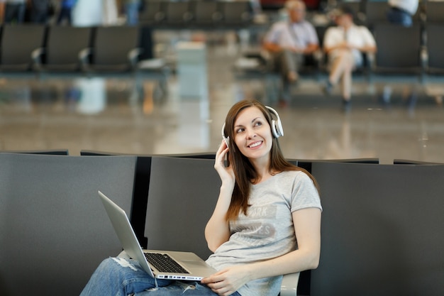 Junge nachdenkliche Reisende Touristenfrau mit Kopfhörern, die Musik hören, die am Laptop arbeitet, warten in der Lobbyhalle am internationalen Flughafen