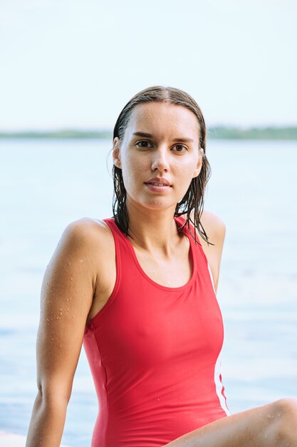 Junge nasse entspannte Frau in rotem Monokini, die vor der Kamera am Wasser sitzt und die Einsamkeit und den Urlaub im Sommerresort genießt