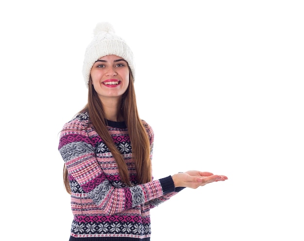 Junge nette Frau im purpurroten Pullover und im weißen Hut, die etwas auf weißem Hintergrund im Studio halten