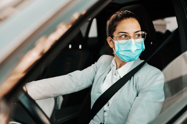 Junge niedliche Brünette gekleidet klug lässig mit Gesichtsmaske beim Fahren ihres Autos während des Ausbruchs des Koronavirus.