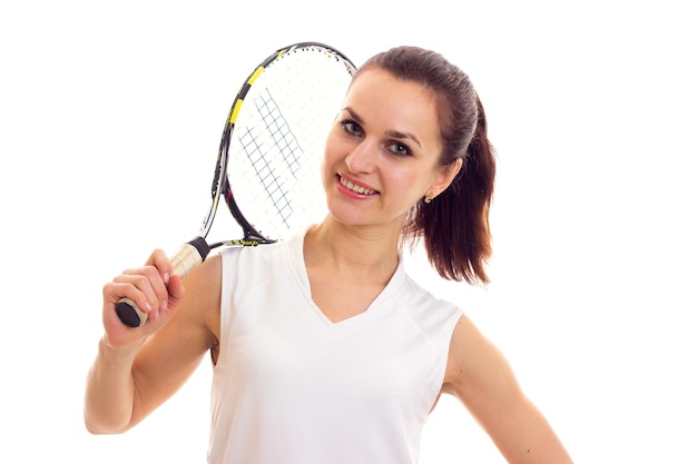 Junge optimistische Frau im weißen Sporthemd mit dunklem Pferdeschwanz, die Tennisschläger im Studio hält