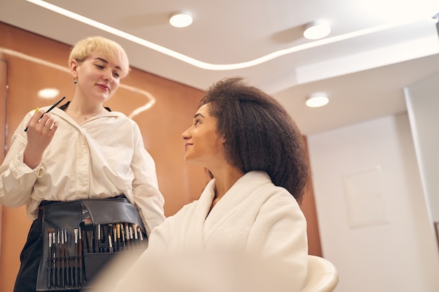 Junge optimistische Visagistin, die ihren charmanten Kunden anschaut, während sie Make-up-Pinsel hält holding