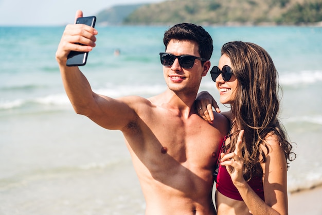 Junge Paare des romantischen Liebhabers, die zusammen am tropischen Strand entspannen. Mann und Frau, die selfie mit Smartphone nehmen und das Leben genießen. Sommerferien