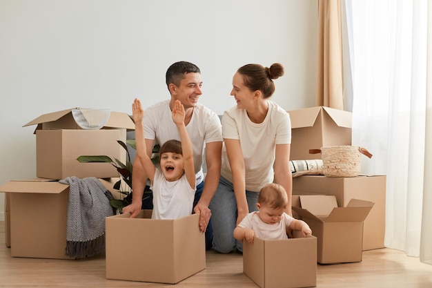 Junge Paare, die in eine neue Wohnung umziehen, ihre Kinder in Kartons reiten, zusammen Faun haben, sich sanft und lächelnd ansehen, den Umzug in ein neues Haus feiern.