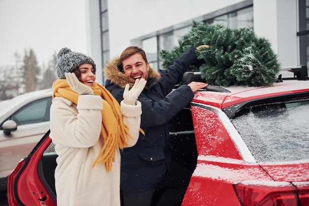 Junge Paare, die nahe Auto mit Baum auf der Oberseite stehen. Zusammen draußen im Winter.