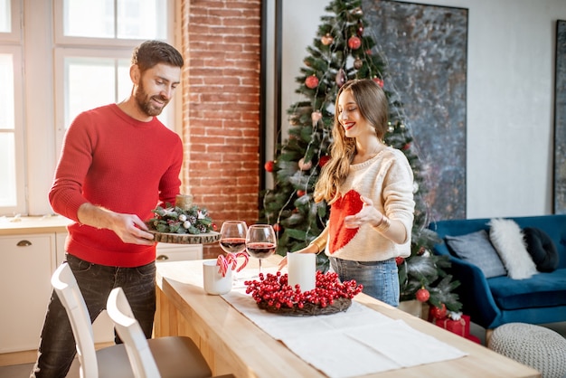 Junge Paare, die sich auf ein neues Jahr vorbereiten und ihre schöne Wohnung im Esszimmer mit Weihnachtsbaum im Hintergrund dekorieren