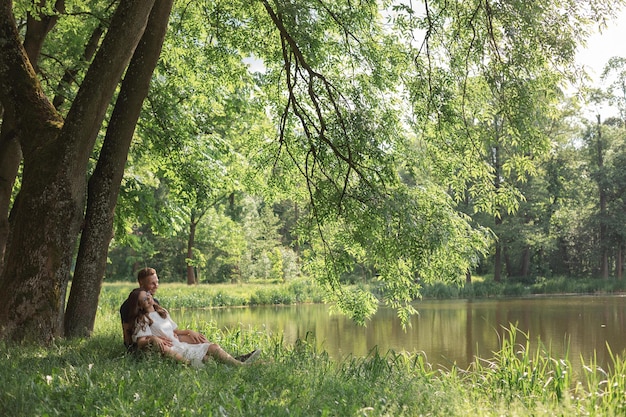 Junge Paare, die sich verlieben, sitzen auf Gras unter einem Baum in der Nähe eines Sees im Park und sehen sich an
