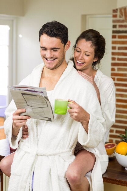 Junge Paare im Bademantel, der Tee trinkt und Zeitung in der Küche liest
