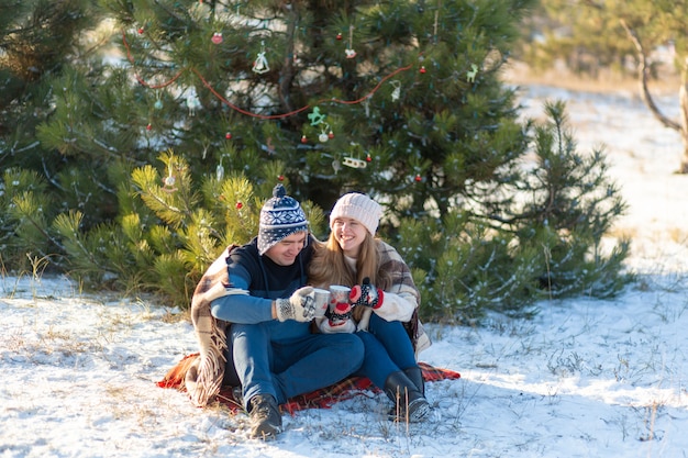 Junge Paare in der Liebe trinken ein heißes Getränk mit den Eibischen und sitzen im Winter im Wald, verstaut in den warmen, bequemen Wolldecken und genießen Natur