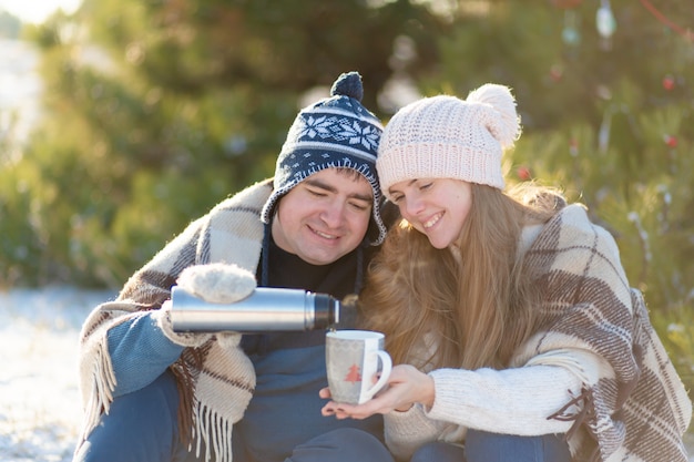 Junge Paare in der Liebe trinken ein heißes Getränk von einer Thermosflasche und sitzen im Winter im Wald, verstaut in warmem