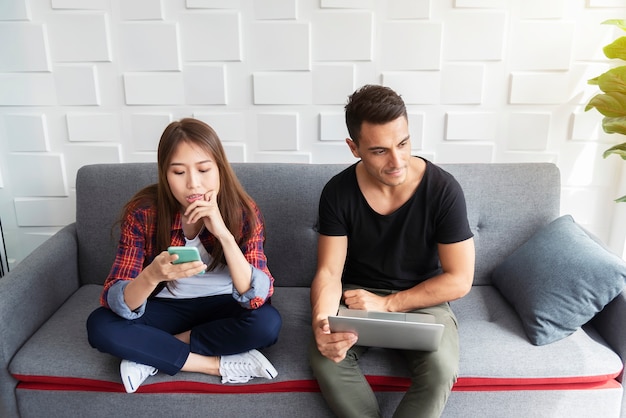 Junge Paare unter Verwendung des Mobiles und des Laptops im Wohnzimmer zu Hause.