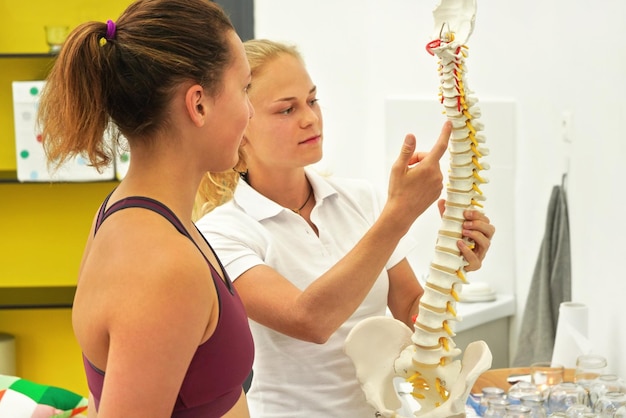 Junge Patientin bei Physiotherapie-Beratung, Physiotherapeutin erklärt Rückenknochen am Wirbelsäulenmodell