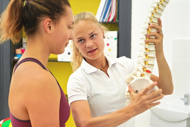 Foto junge patientin bei physiotherapie-beratung, physiotherapeutin erklärt rückenknochen am wirbelsäulenmodell