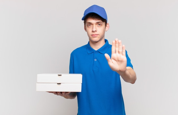 Junge Pizza liefern Jungen, die ernst, streng, unzufrieden und wütend aussehen und eine offene Handfläche zeigen, die eine Stopp-Geste macht