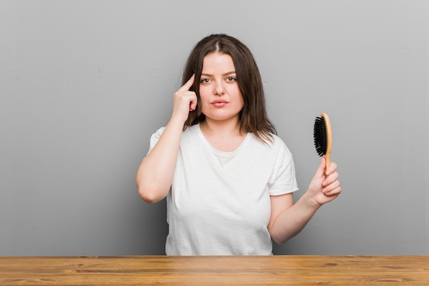 Junge plus die curvy Frau der Größe, die eine Haarbürste zeigt seinen Tempel mit dem Finger, denkend anhält, gerichtet auf eine Aufgabe.