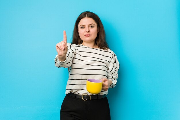 Junge plus Größe Frau, die eine Teetasse hält, die Nummer eins mit Finger zeigt.