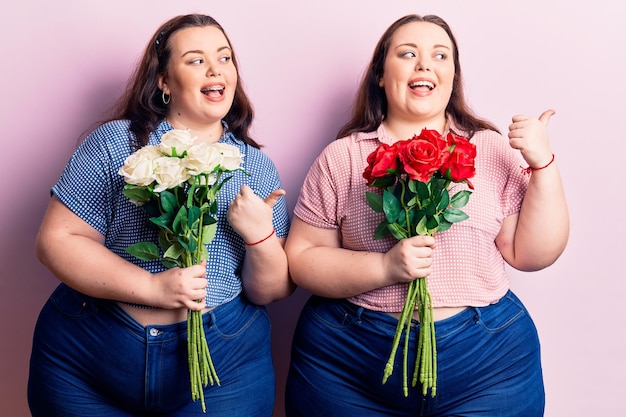 Junge Plus-Size-Zwillinge mit Blumen, die mit dem Daumen nach oben zur Seite zeigen und glücklich mit offenem Mund lächeln