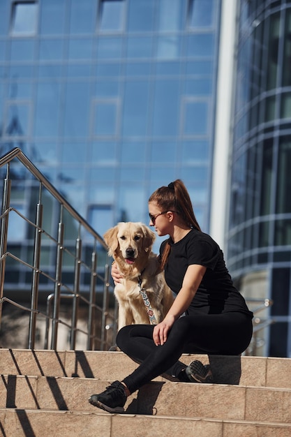 Junge positive Frau, die mit ihrem Hund auf der Treppe sitzt, wenn sie draußen in der Nähe des Geschäftsgebäudes spazieren geht