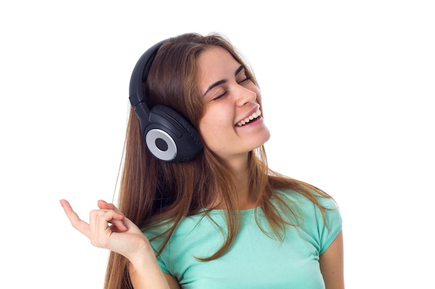 Junge positive Frau im blauen T-Shirt, die Musik in schwarzen Kopfhörern mit geschlossenen Augen auf weißem Hintergrund im Studio hört