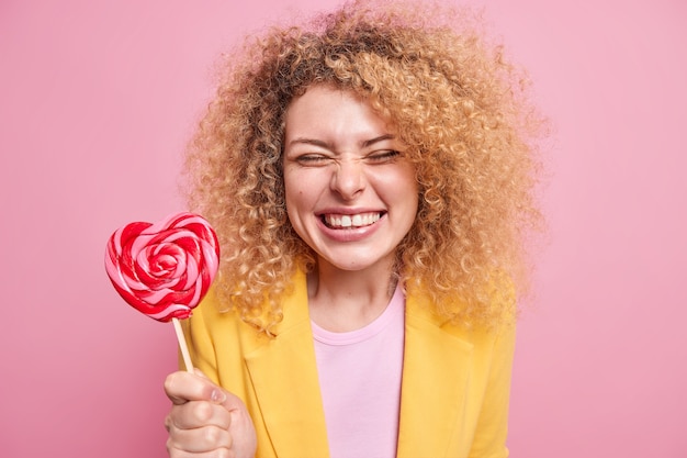 Junge positive Frau mit blonden Locken hält herzförmige Karamellbonbons und lächelt zähnefletschend