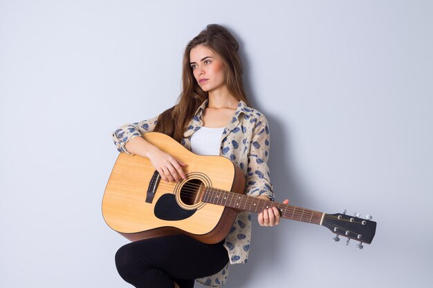 Junge positive Frau mit langen Haaren in brauner Jacke, die im Studio Gitarre spielt