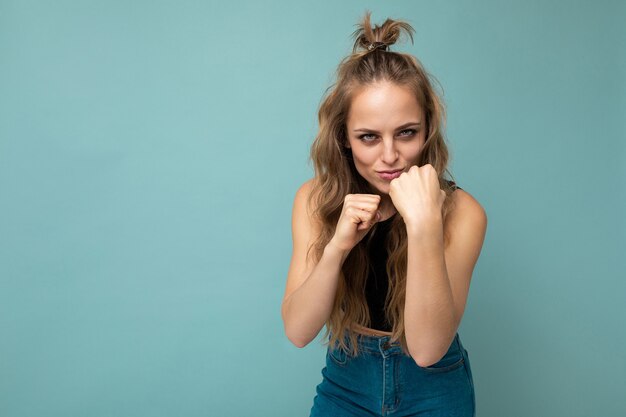 Junge positive konzentrierte schöne blonde blonde wellige Frau mit aufrichtigen Gefühlen, die trendiges schwarzes Oberteil lokalisiert über blauem Hintergrund mit leerem Raum tragen und bereit sind zu kämpfen.