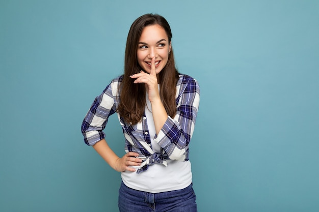 Junge positive lächelnde schöne Brunetfrau mit aufrichtigen Gefühlen, die trendiges Karohemd tragen, das lokal auf Blau steht