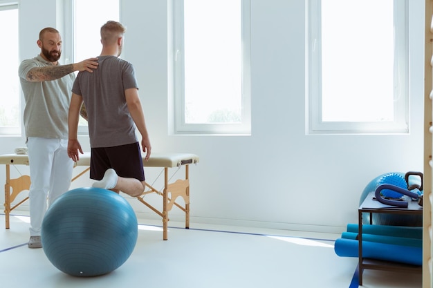 Junge professionelle Physiotherapeutin trainiert mit einem verletzten Patienten mit einem blauen Ball