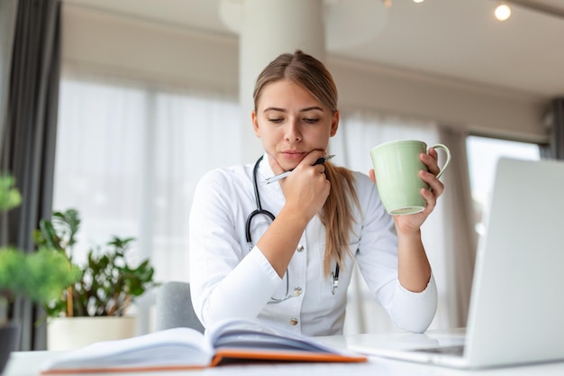 Junge professionelle Ärztin in Uniform, die sich Notizen in einem medizinischen Journal macht, das Dokumente über die Krankheitsgeschichte des Patienten ausfüllt und auf den Laptop-Bildschirm schaut, Student, der sich ein Webinar ansieht