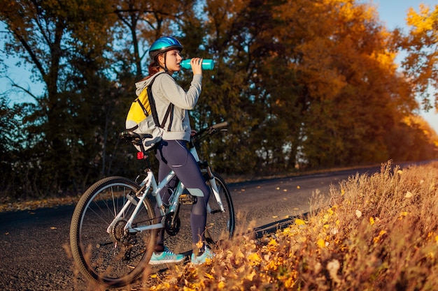 Junge Radfahrer, die sich nach einer Fahrt im Herbstfeld bei Sonnenuntergang ausruhen. Trinkwasser der Frau auf der Straße. Gesunder Lebensstil