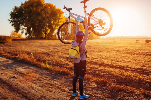 Junge Radfahrerin, die bei Sonnenuntergang ihr Fahrrad im Herbstfeld anhebt. Glückliche Frau feiert den Sieg, der Fahrrad in den Händen hält.