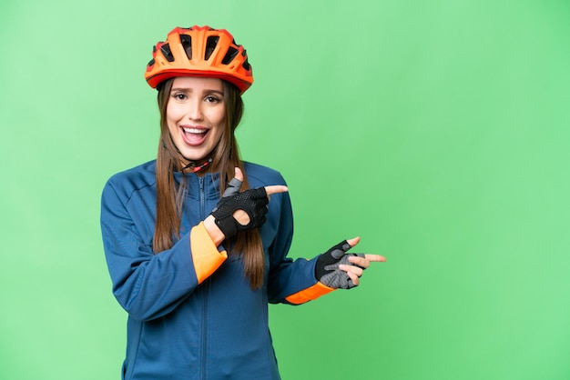 Junge Radfahrerin vor isoliertem Chroma-Key-Hintergrund überrascht und zeigt zur Seite