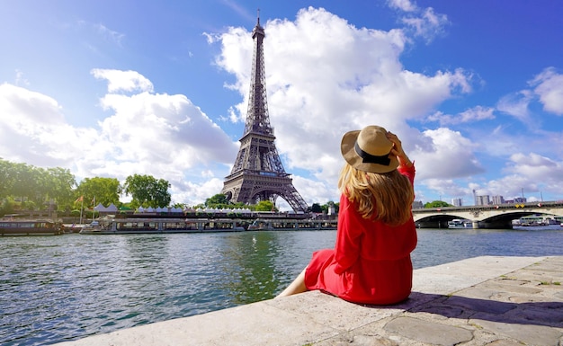 Junge Reisende Frau in rotem Kleid und Hut sitzt am Kai der Seine und blickt auf das berühmte Wahrzeichen und Reiseziel des Eiffelturms in Paris