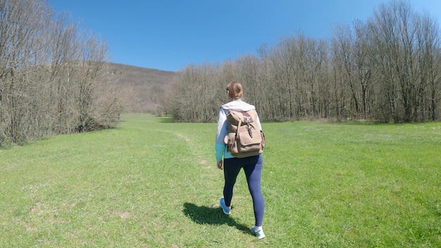 Junge Reisende mit Rucksack geht den Weg entlang, vorbei an Wiesen und Wäldern. Konzept für Reisen und Outdoor-Aktivitäten. Rückansicht. 4K UHD