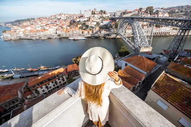 Junge Reisende mit Sonnenhut, die während des Morgenlichts in Porto, Portugal, einen schönen Blick auf das Stadtbild mit dem Fluss Douro und der Luise-Brücke genießen