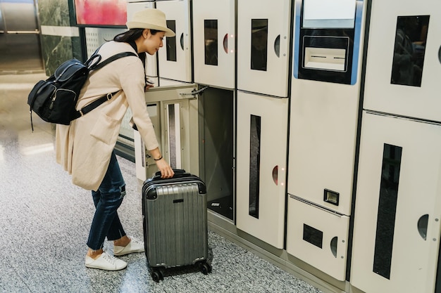 junge reisende rucksacktouristin, die gepäck in münzfach im lobby-flughafen legt. Starke, elegante Touristenfrau mit Hut, die schwere Koffer aufnimmt, öffnet die Tür. reise in den frühlingsferien.