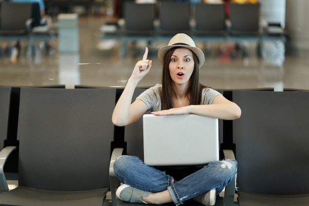 Junge reisende Touristenfrau mit Laptop, die mit gekreuzten Beinen sitzt, den Finger hochhält und Idee hat und in der Lobbyhalle am Flughafen wartet