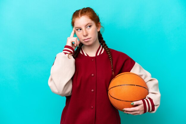 Junge rötliche Frau, die Basketball spielt, isoliert auf blauem Hintergrund, hat Zweifel und denkt nach