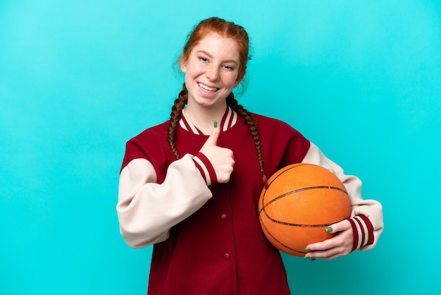 Junge rötliche Frau spielt Basketball isoliert auf blauem Hintergrund und gibt eine Daumen hoch Geste