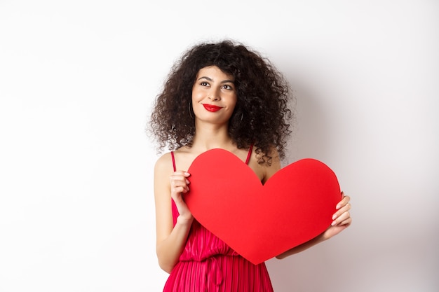 Junge romantische Frau träumt von Liebe am Valentinstag, sucht Seelenverwandten, hält großen roten Herzausschnitt und schaut nachdenklich links, weißer Hintergrund.