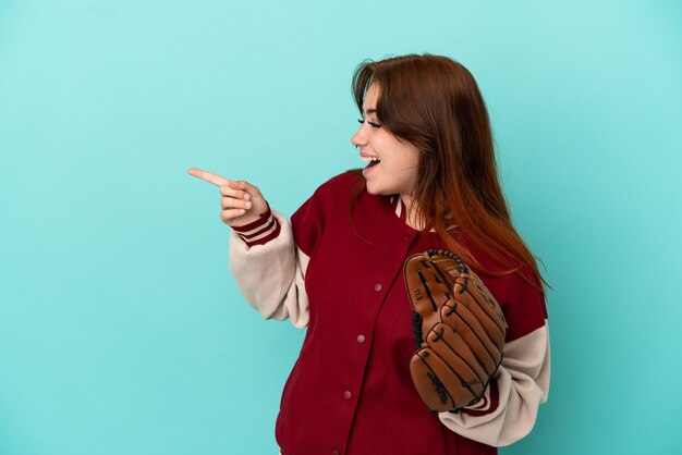 Junge rothaarige Frau, die Baseball spielt, isoliert auf blauem Hintergrund, der mit dem Finger zur Seite zeigt und ein Produkt präsentiert