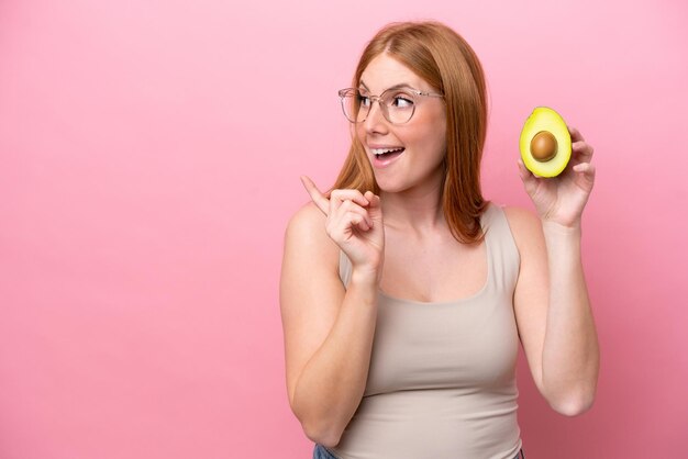 Junge rothaarige Frau, die eine Avocado isoliert auf rosa Hintergrund hält und beabsichtigt, die Lösung zu realisieren, während sie einen Finger hochhebt