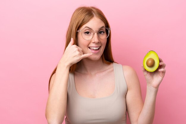 Junge rothaarige Frau, die eine Avocado isoliert auf rosafarbenem Hintergrund hält und eine Telefongeste macht Ruf mich zurück