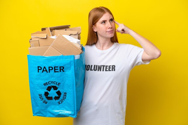 Junge rothaarige Frau, die eine Recyclingtüte voller Papier hält, um isoliert auf gelbem Hintergrund zu recyceln, die Zweifel hat und nachdenkt