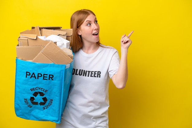 Junge rothaarige Frau, die eine Recyclingtüte voller Papier hält, um sie isoliert auf gelbem Hintergrund zu recyceln, um die Lösung zu realisieren, während sie einen Finger hochhebt