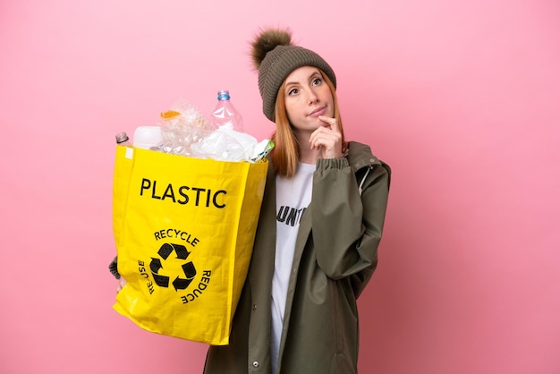 Junge rothaarige Frau, die eine Tüte voller Plastikflaschen hält, um sie isoliert auf rosafarbenem Hintergrund zu recyceln und nach oben zu schauen