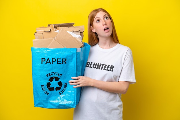 Junge rothaarige Frau, die einen Recycling-Beutel voller Papier hält, um isoliert auf gelbem Hintergrund zu recyceln und nach oben zu schauen und mit überraschtem Ausdruck