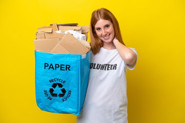 Junge rothaarige Frau, die einen Recycling-Beutel voller Papier hält, um isoliert auf gelbem Hintergrund zu recyceln und zu lachen