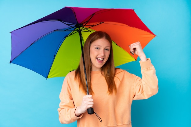 Junge rothaarige Frau, die einen Regenschirm über isolierter Wand hält, die einen Sieg feiert