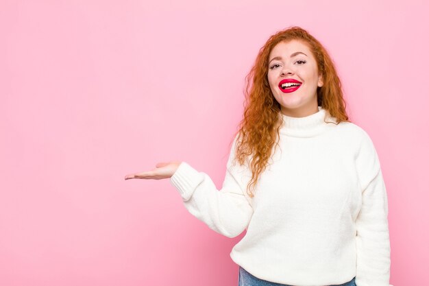 Junge rothaarige Frau, die fröhlich lächelt, sich glücklich fühlt und ein Konzept im Kopierraum mit Handfläche gegen rosa Wand zeigt